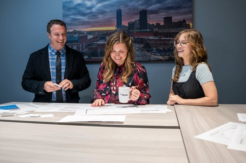 Three colleagues stand around a conference table reviewing papers; the person in the center holds a coffee mug while the others look on and smile.